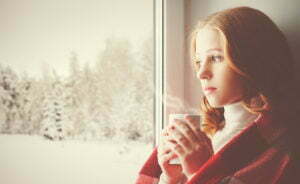 Pensive sad girl with a warming drink looking out the window in the winter forest