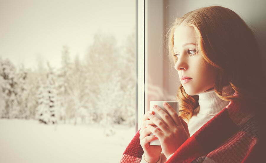 Pensive sad girl with a warming drink looking out the window in the winter forest